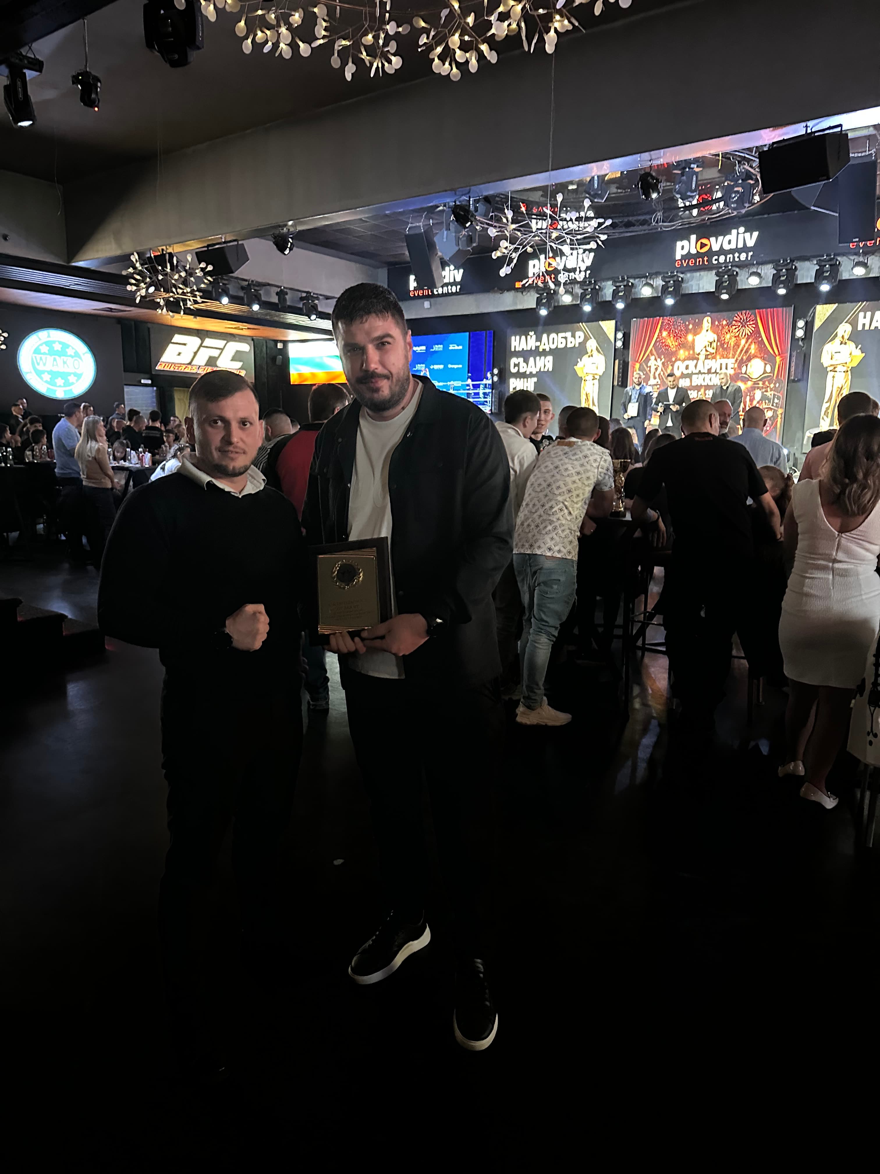 Two men posing with a gold award plaque at a crowded event center with stage lights.