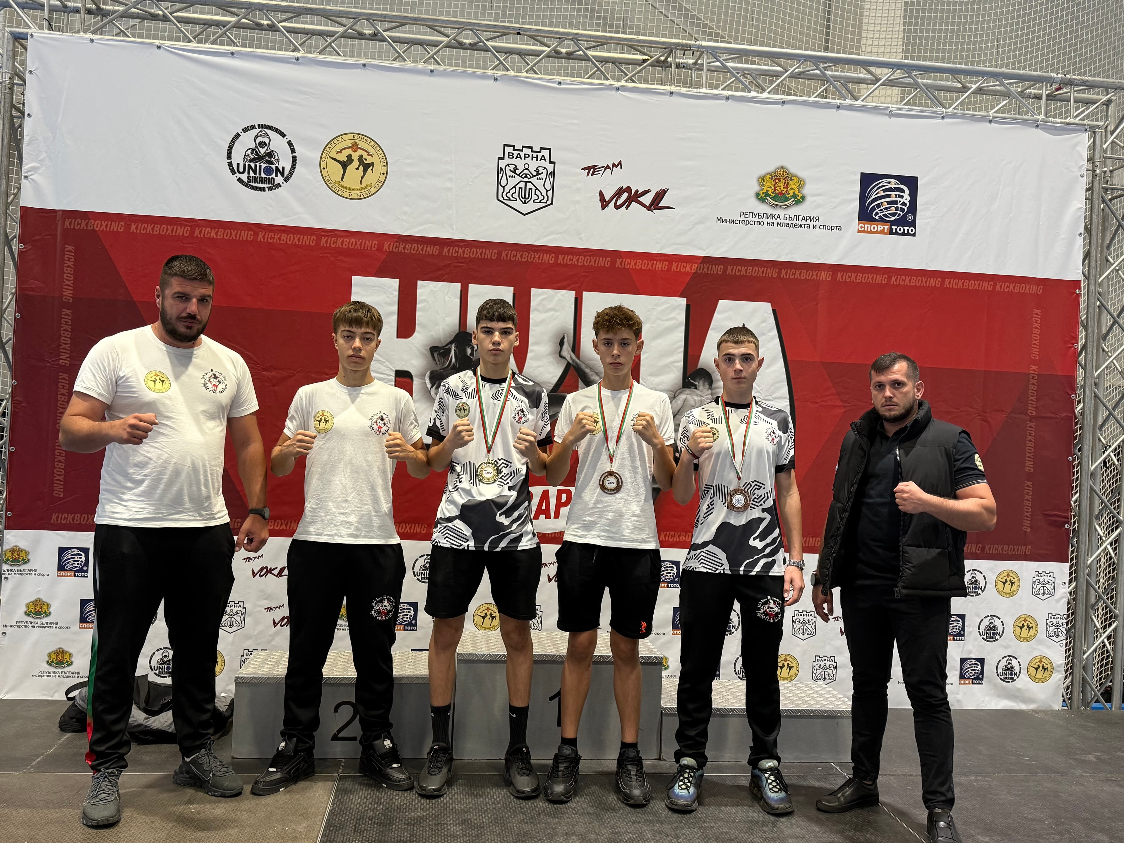 Four young kickboxers with medals pose with two coaches before a large event banner.