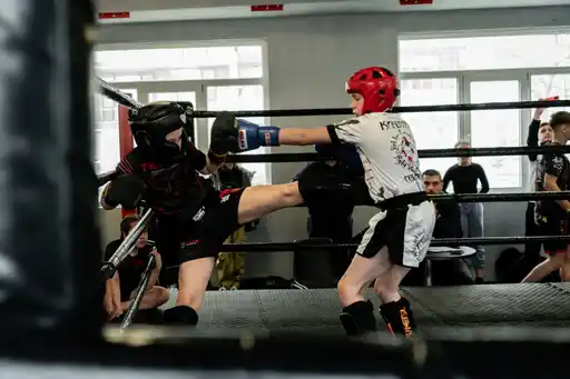 Two young kickboxers in protective gear sparring in a ring, one landing a kick.