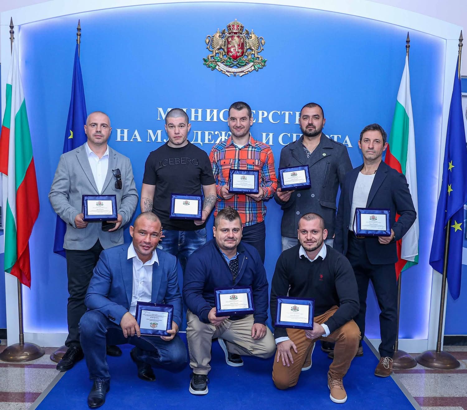 Eight men holding award plaques pose in front of a blue wall with Bulgarian flags.