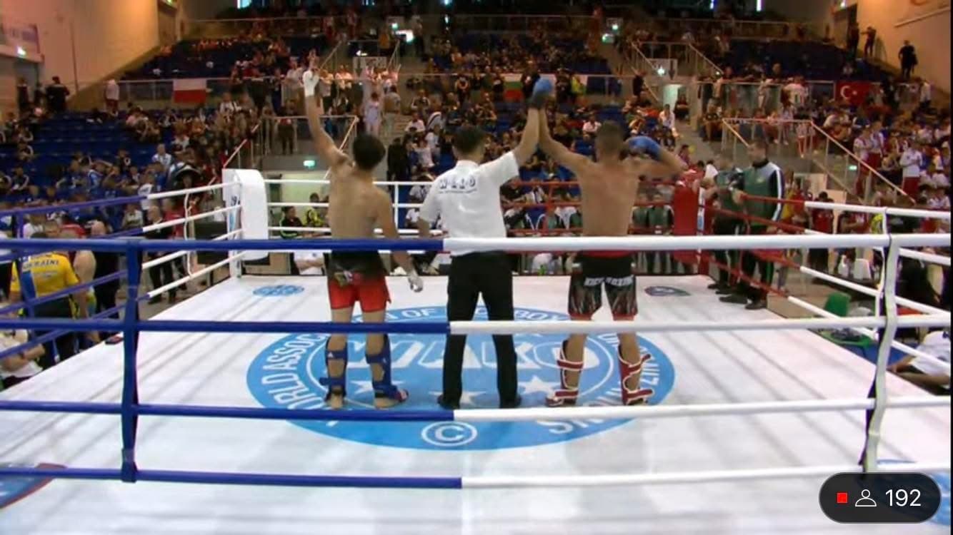 Referee raises the arms of two kickboxers in a ring facing a packed arena.