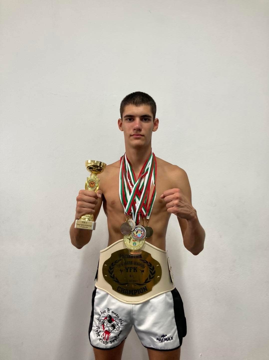 Shirtless young fighter posing with a gold trophy, multiple medals, and a championship belt.