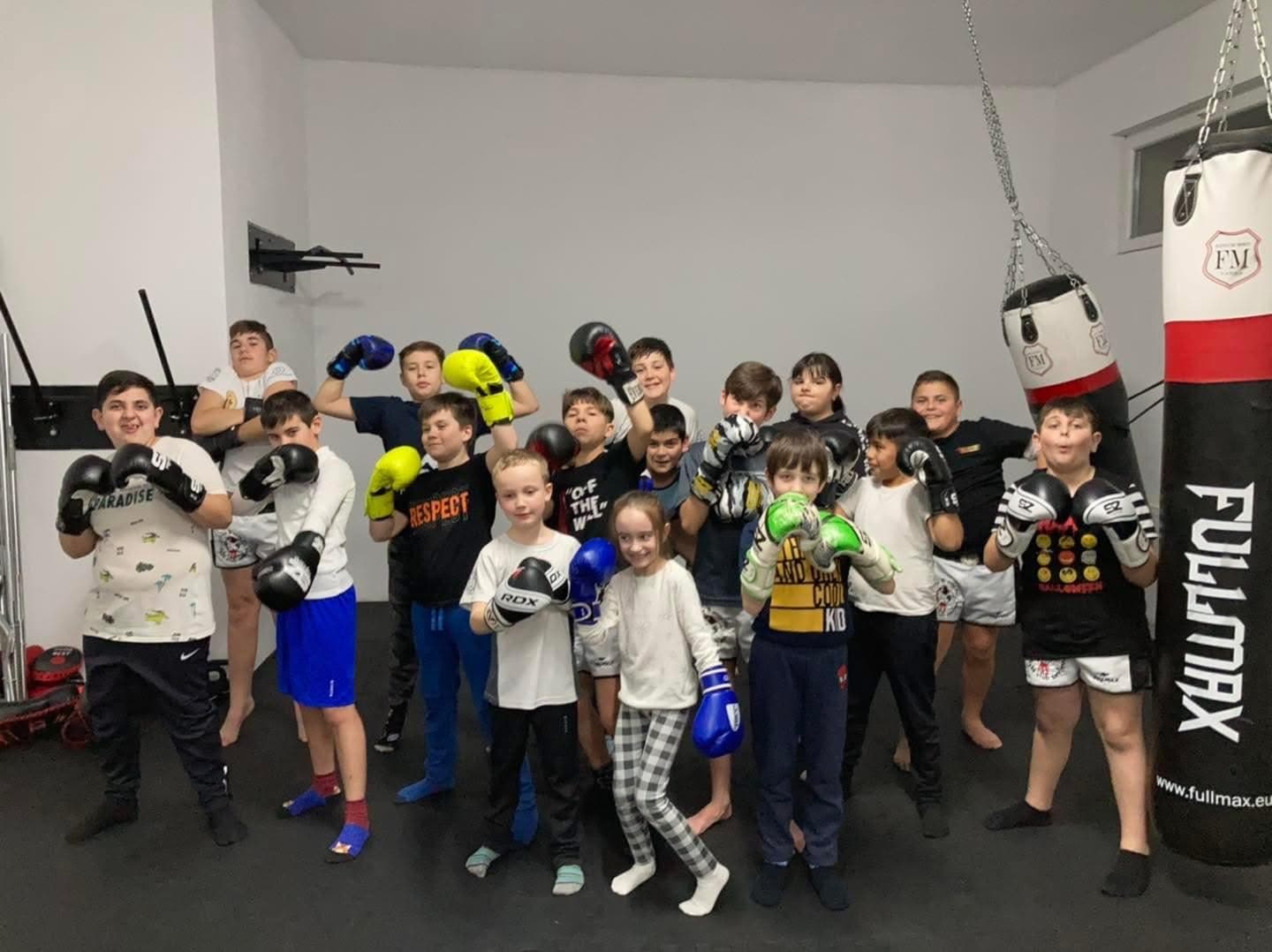A group of children wearing boxing gloves pose in a gym with heavy punching bags.