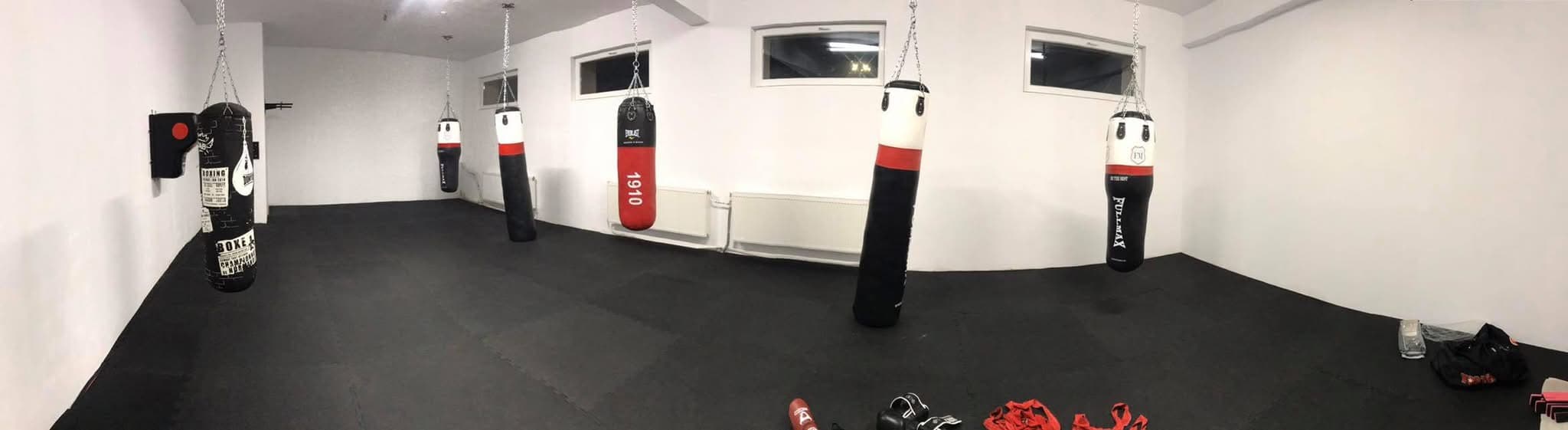Boxing gym featuring several heavy bags hanging over black mats and white walls.