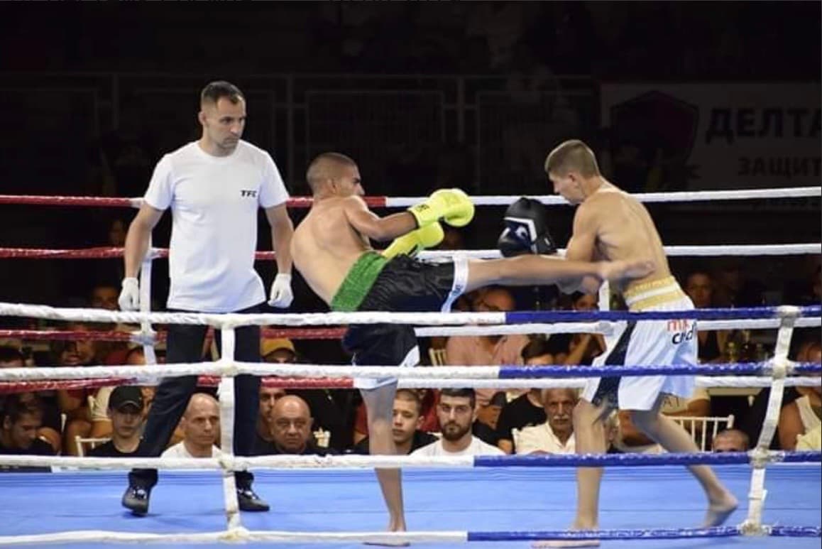 Kickboxer in black shorts delivers a high kick to his opponent while the referee watches.