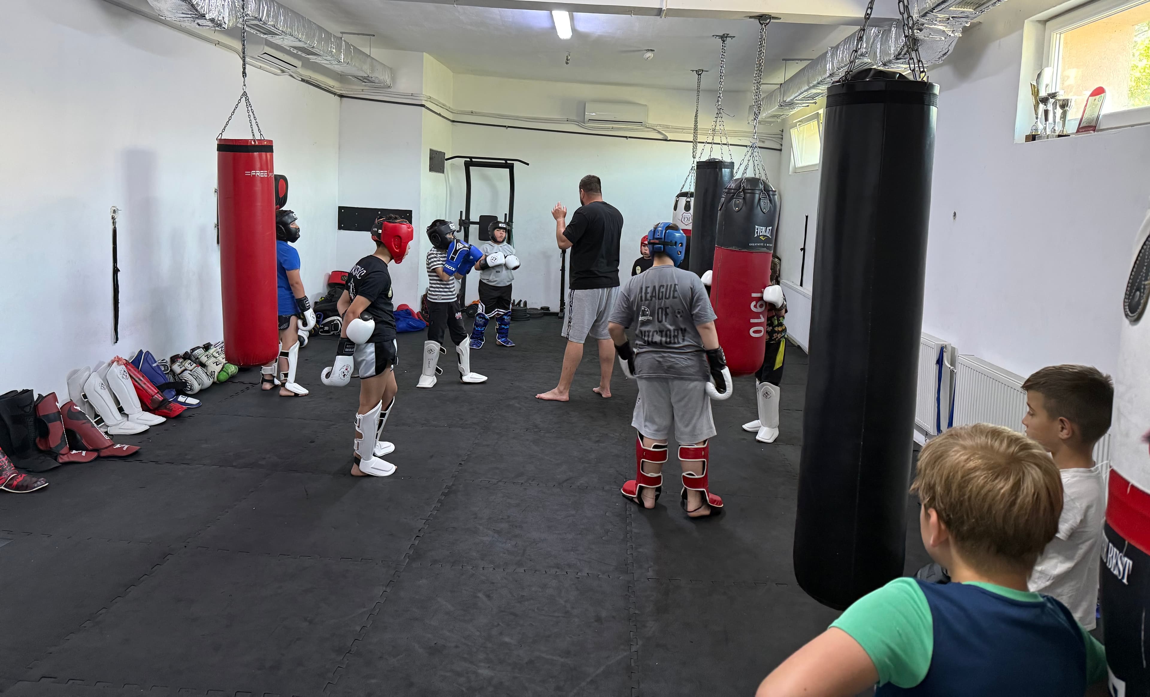 Children wearing protective gear train in a kickboxing gym with a coach and punching bags.