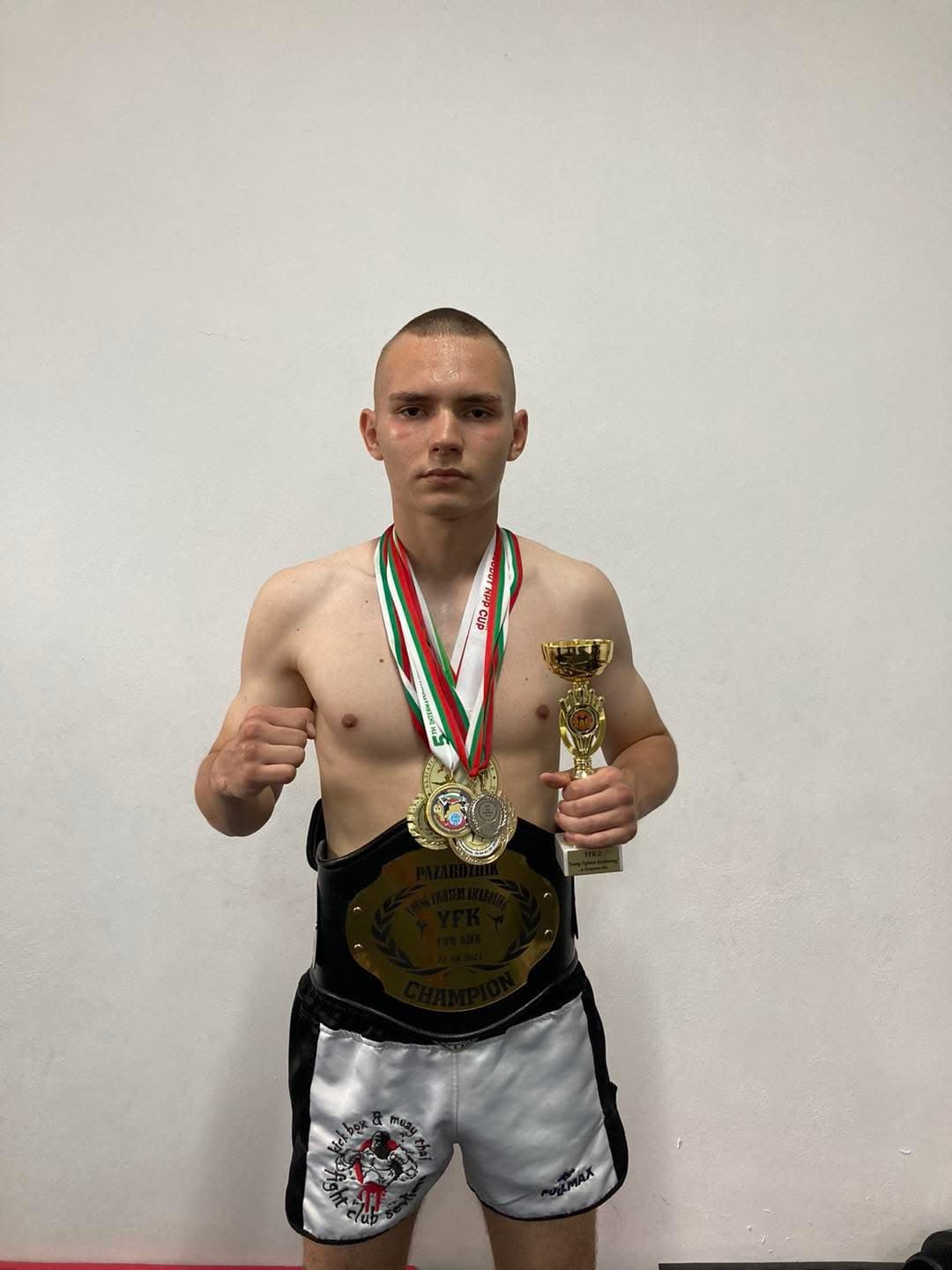 Young shirtless kickboxer wearing a championship belt and medals, holding a trophy against white background.